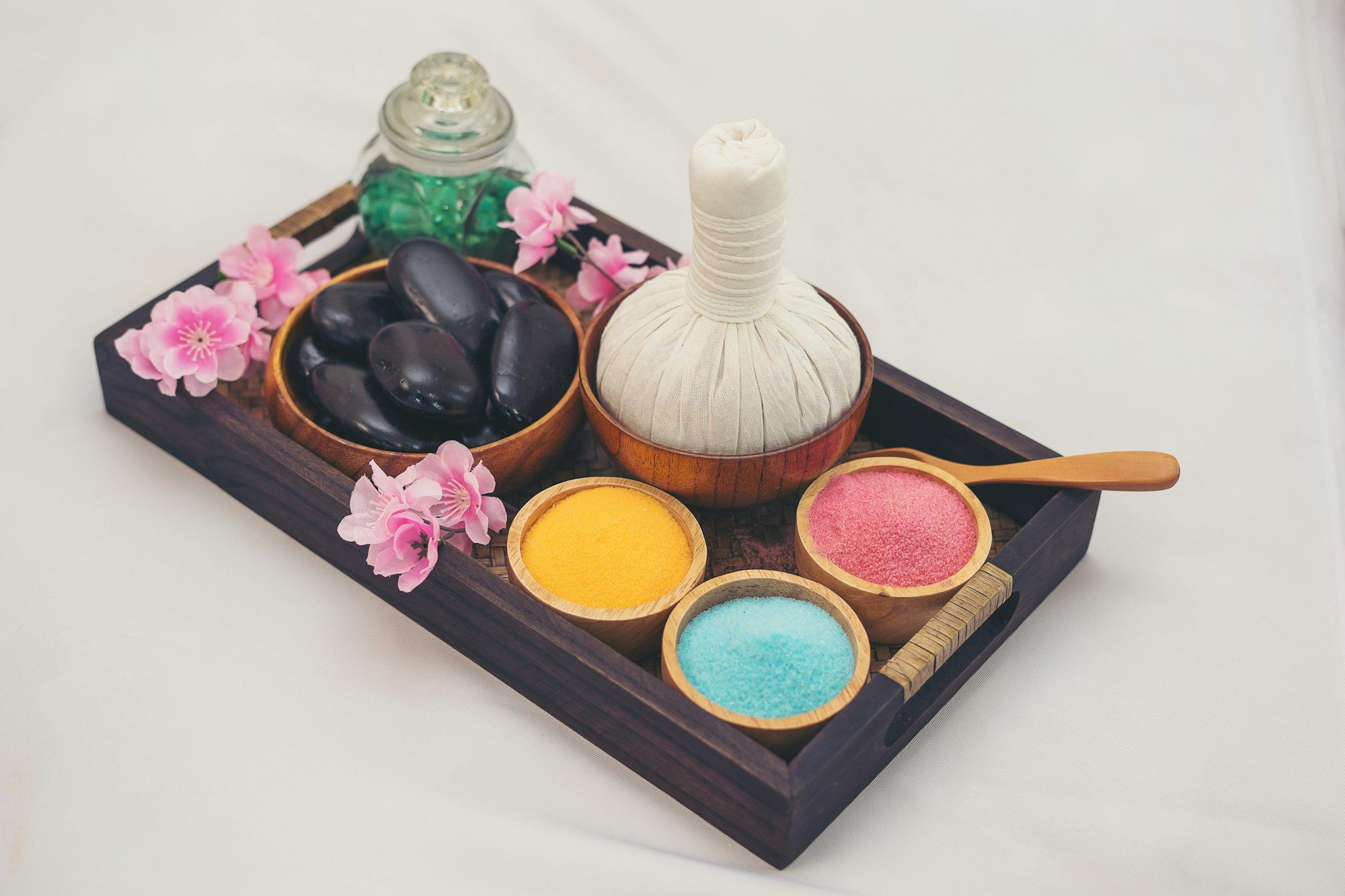 A wooden tray with a herbal poultice bundle, warm stones, coloured salts and pink flowers ready for therapy.