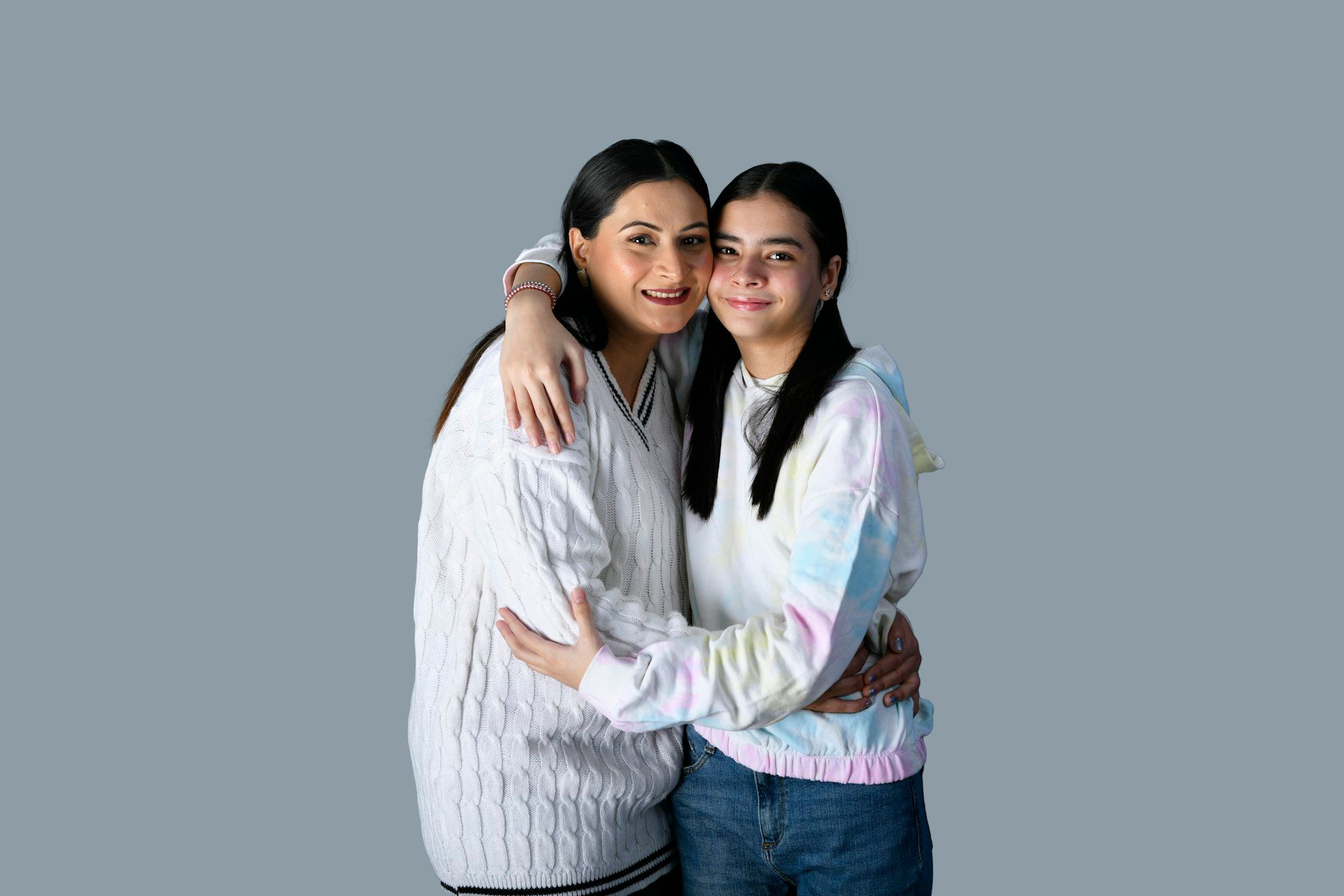An Indian mother and her teenage daughter hugging and smiling against a soft grey backdrop.