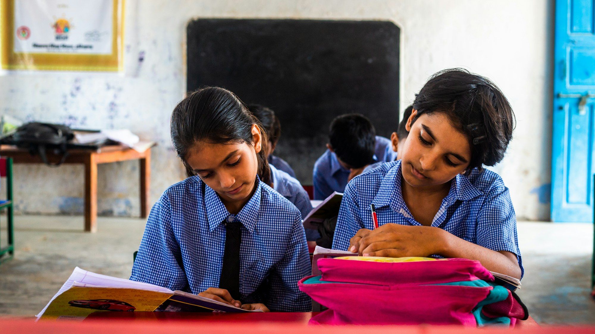 Two Indian schoolgirls in uniform reading and writing at their desk in a classroom.