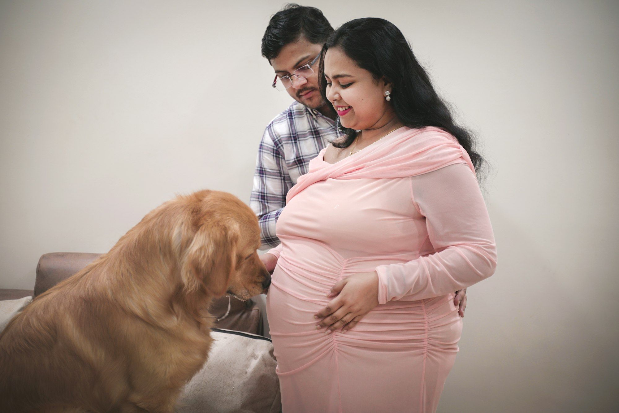 A smiling Indian mother-to-be in a long-sleeved pink dress stands at home with her husband as their golden retriever leans in.