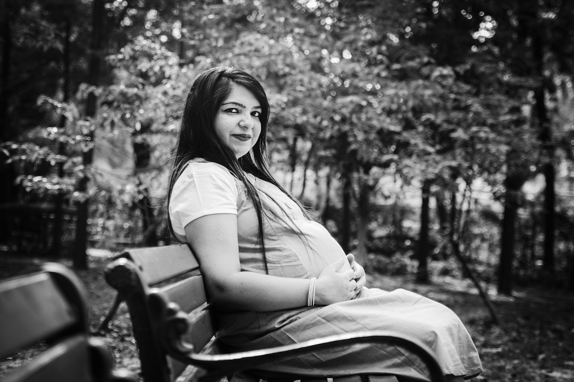 A black-and-white portrait of a smiling Indian mother-to-be sitting on a park bench with her hands on her bump.