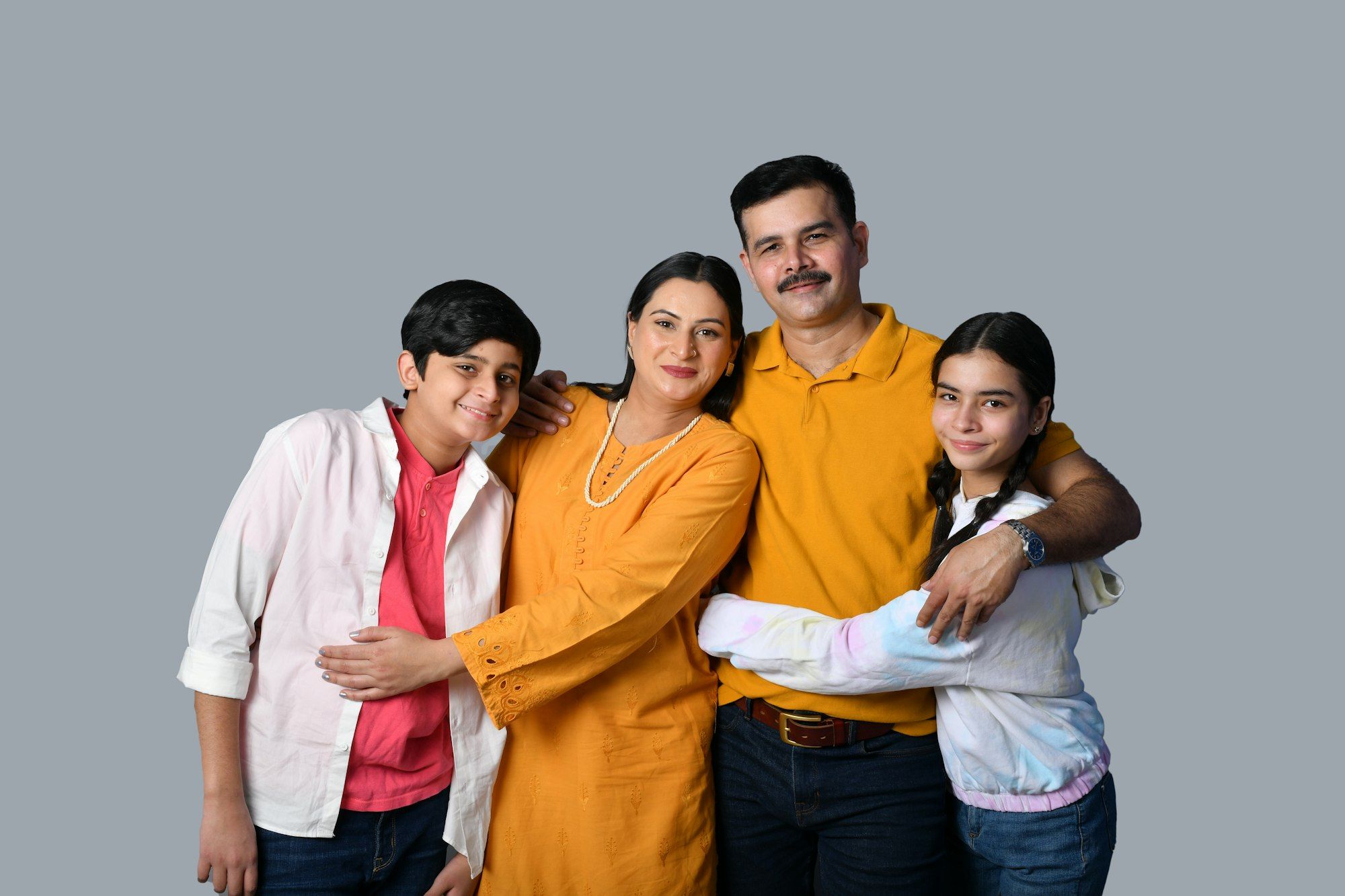 An Indian mother and father smiling with their son and daughter in a warm family group hug against a grey backdrop.