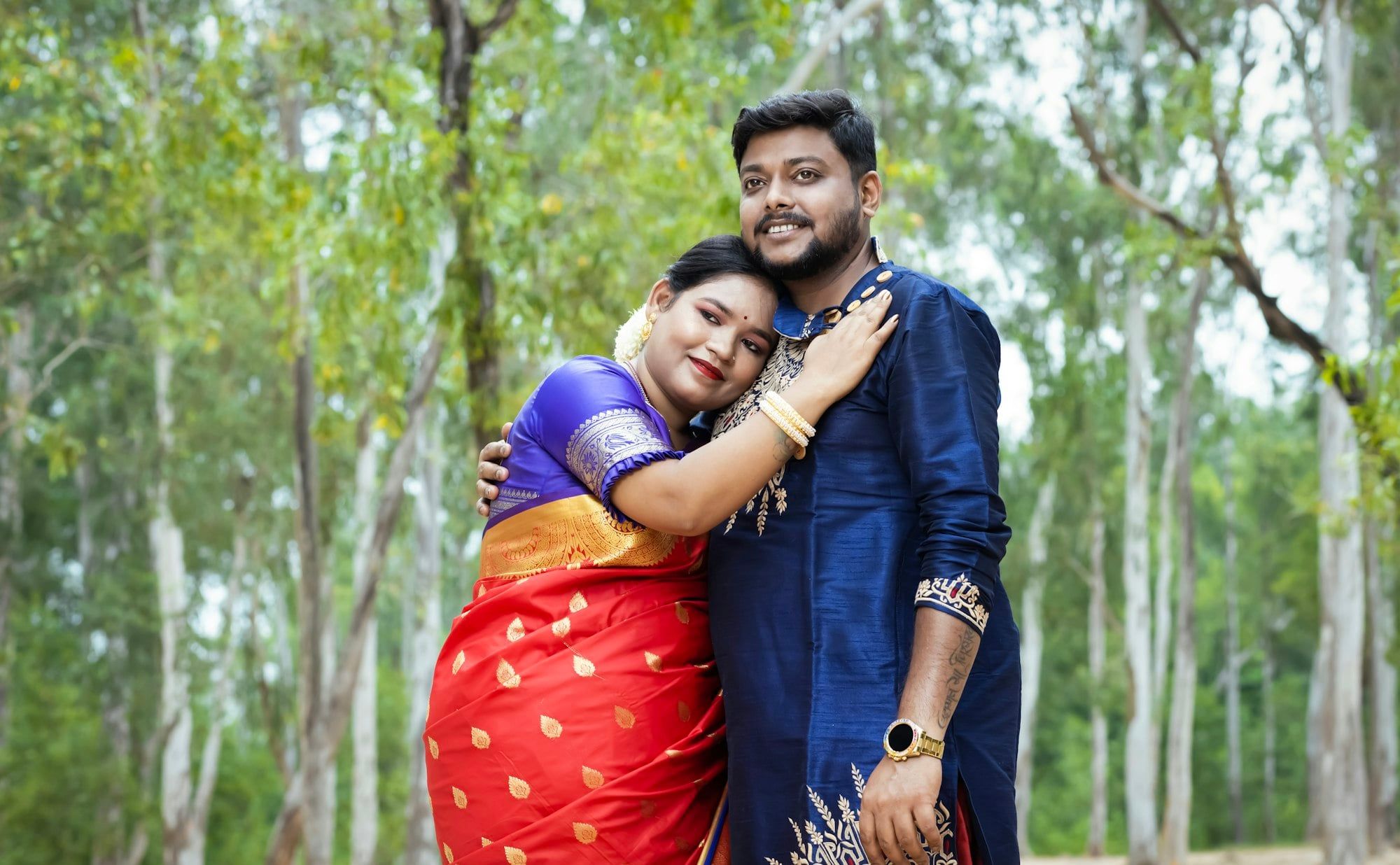 A hopeful Indian couple, she in a red silk saree and he in a navy kurta, share a warm embrace among tall trees.