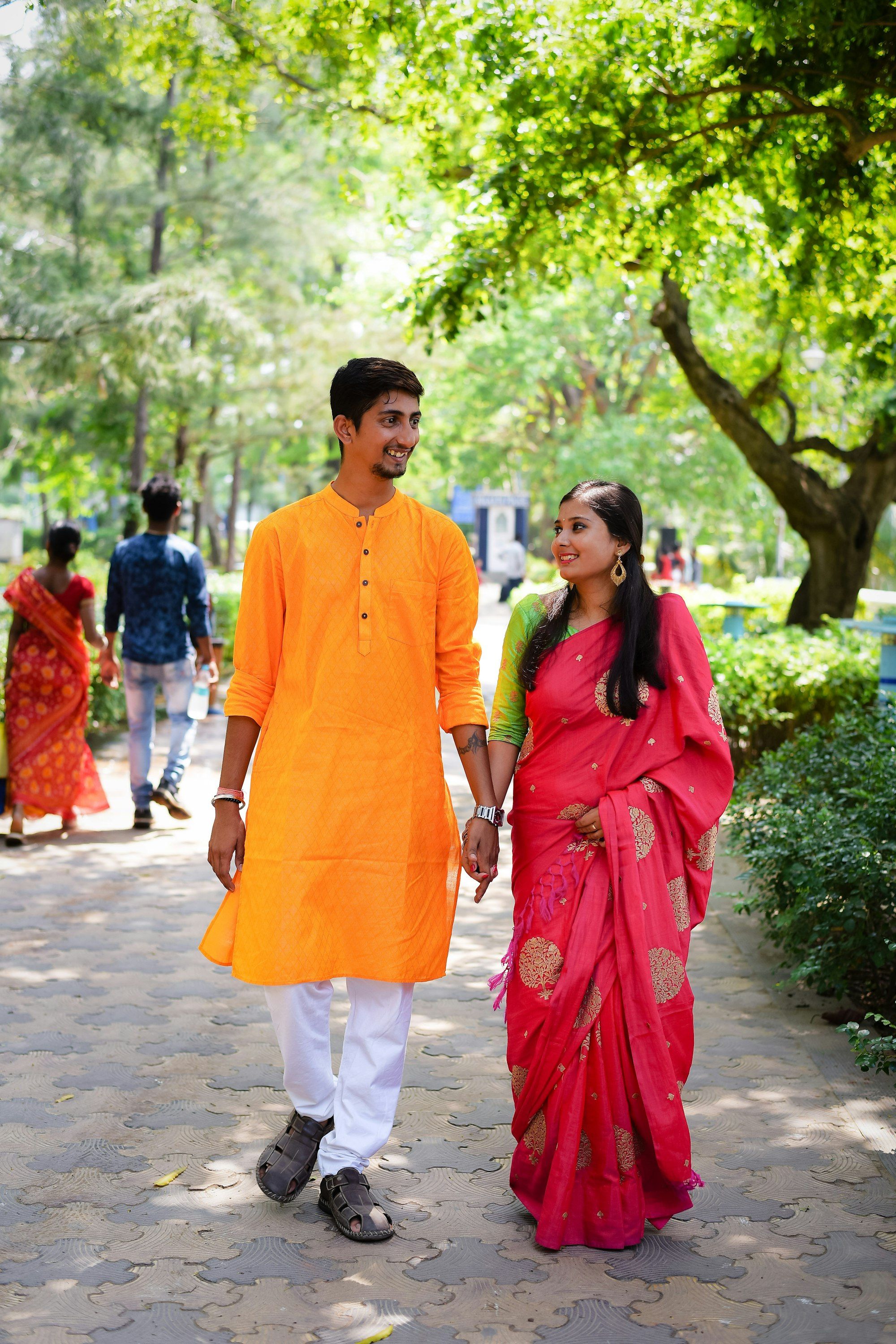 A smiling young Indian couple in a pink saree and saffron kurta walk hand in hand along a leafy park path.