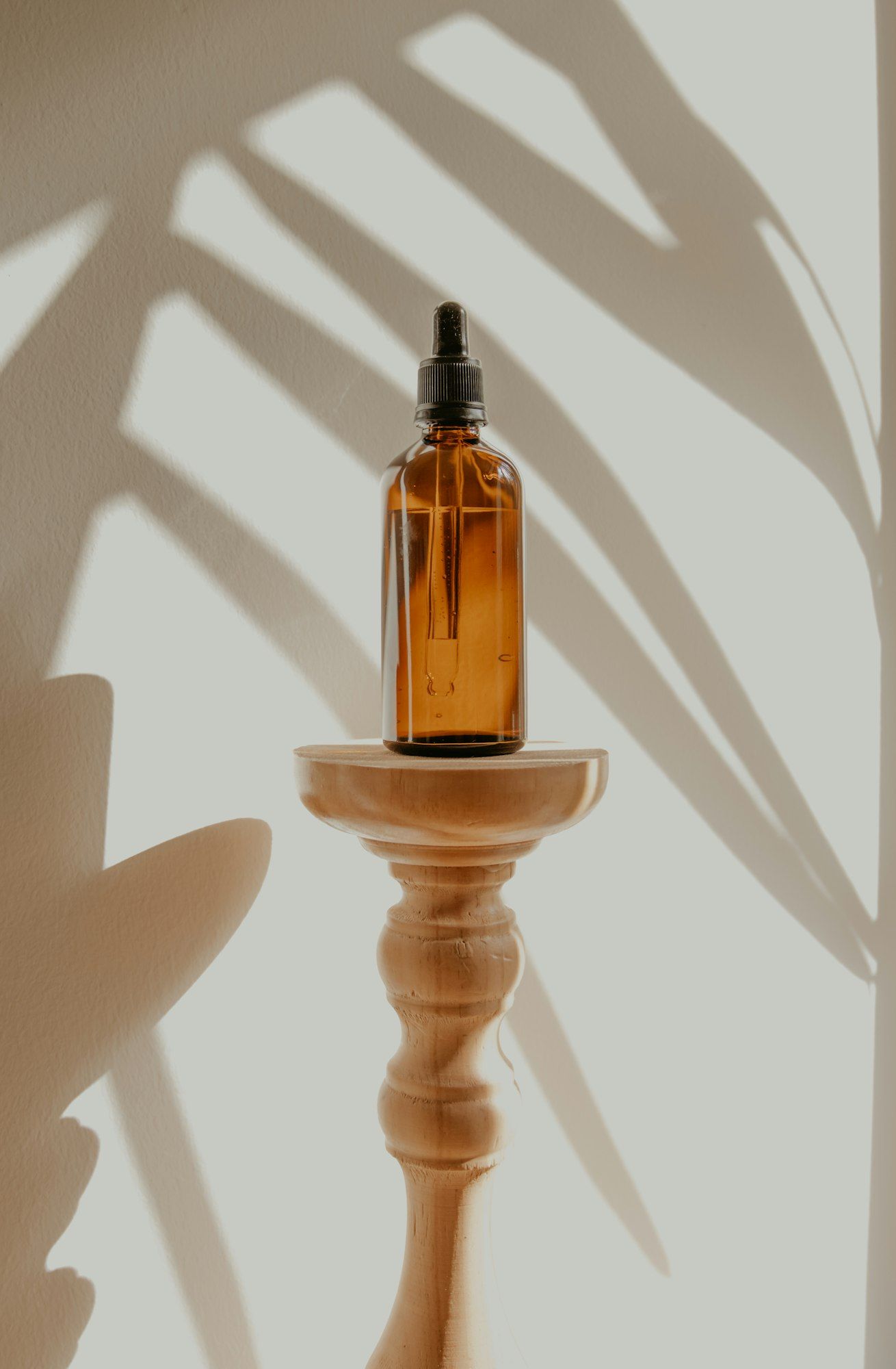 An amber glass dropper bottle of oil on a turned wooden stand with soft leaf shadows behind.