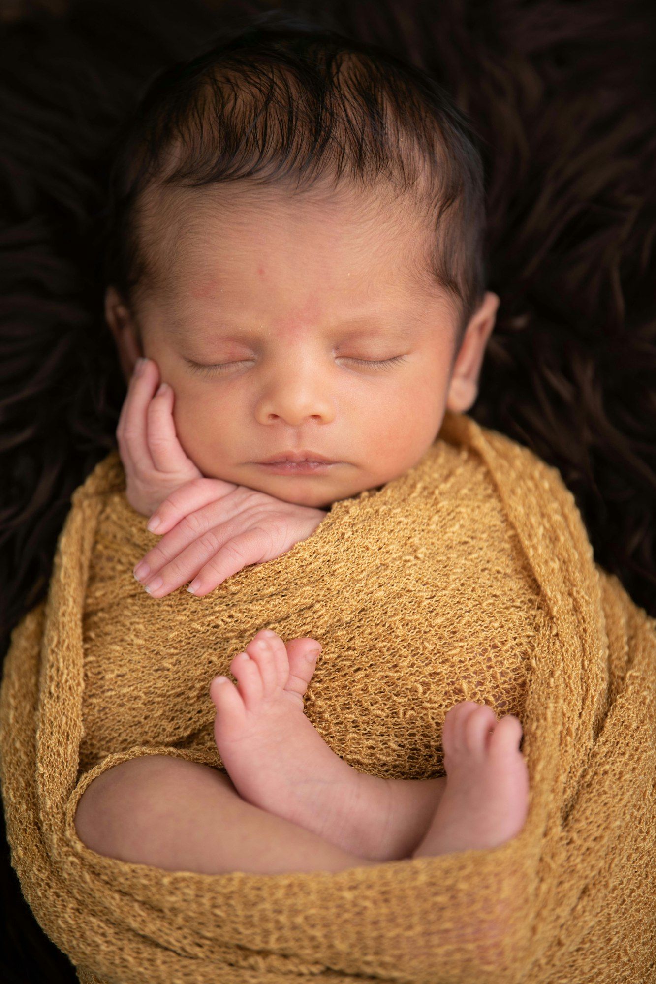 A sleeping newborn baby swaddled in a mustard-yellow wrap with tiny hands resting under the chin.