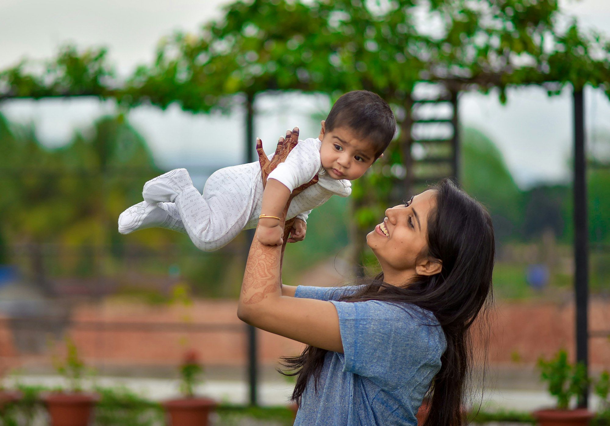 A joyful Indian mother lifting her baby high in the air in a leafy garden.