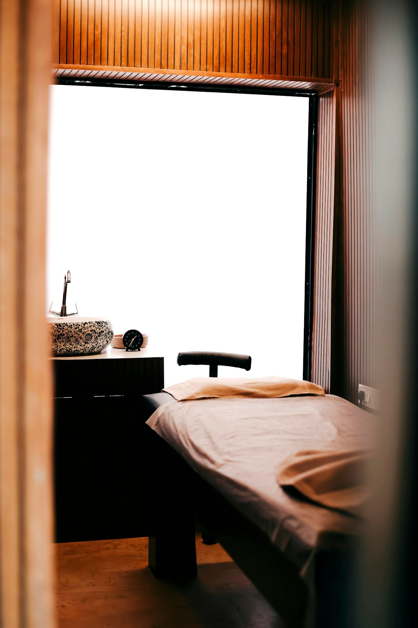 A quiet, warm-toned therapy room with a draped treatment table beside a stone wash basin.