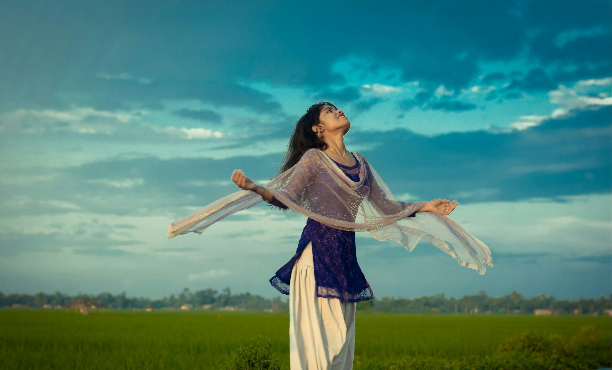 A young Indian woman in a salwar kurta lets her dupatta flow as she breathes in the fresh air over green fields.