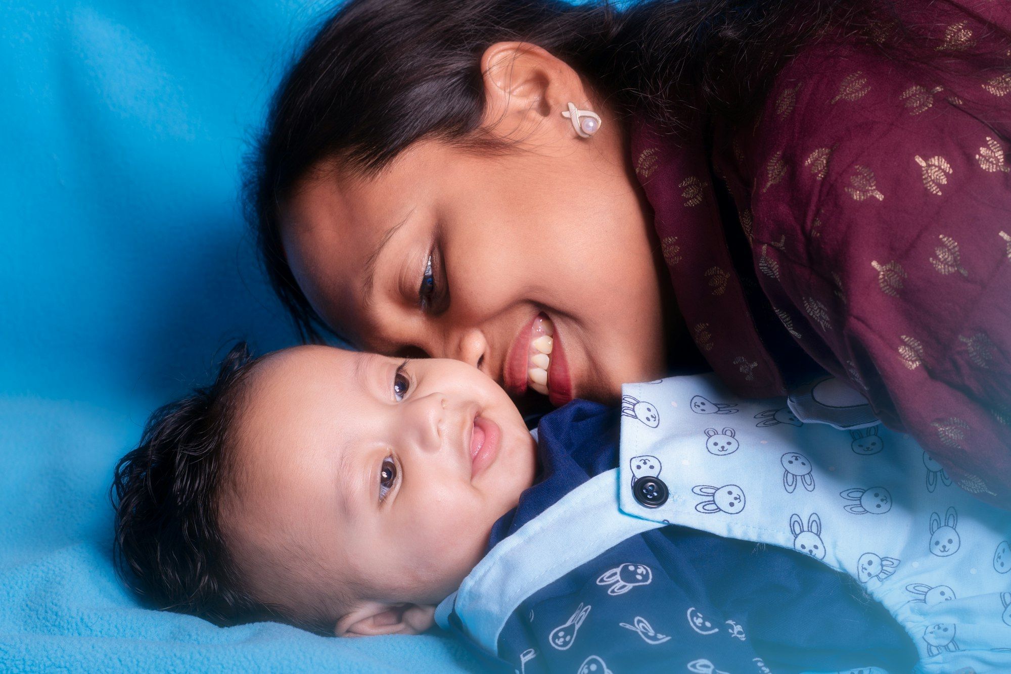 A smiling Indian mother nuzzling her happy baby on a blue background.