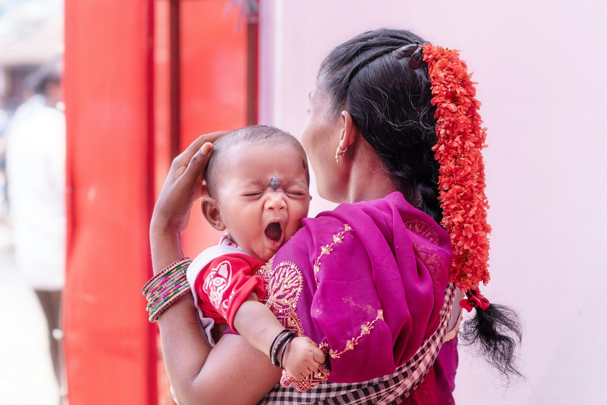 An Indian mother in a pink saree with flowers in her braid cradling her yawning baby.