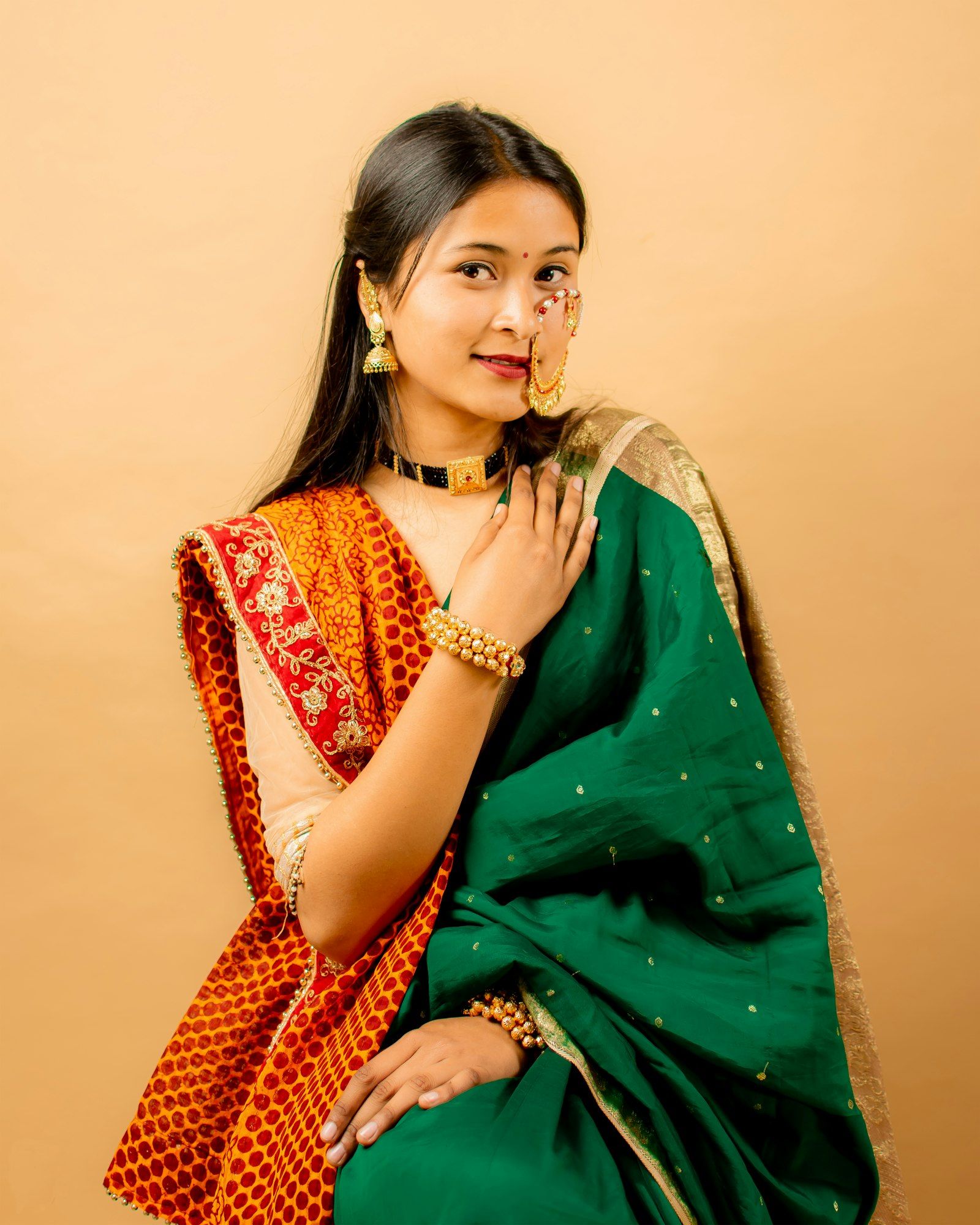 A smiling young Indian woman in a green and red saree with traditional jewellery against a warm peach backdrop.