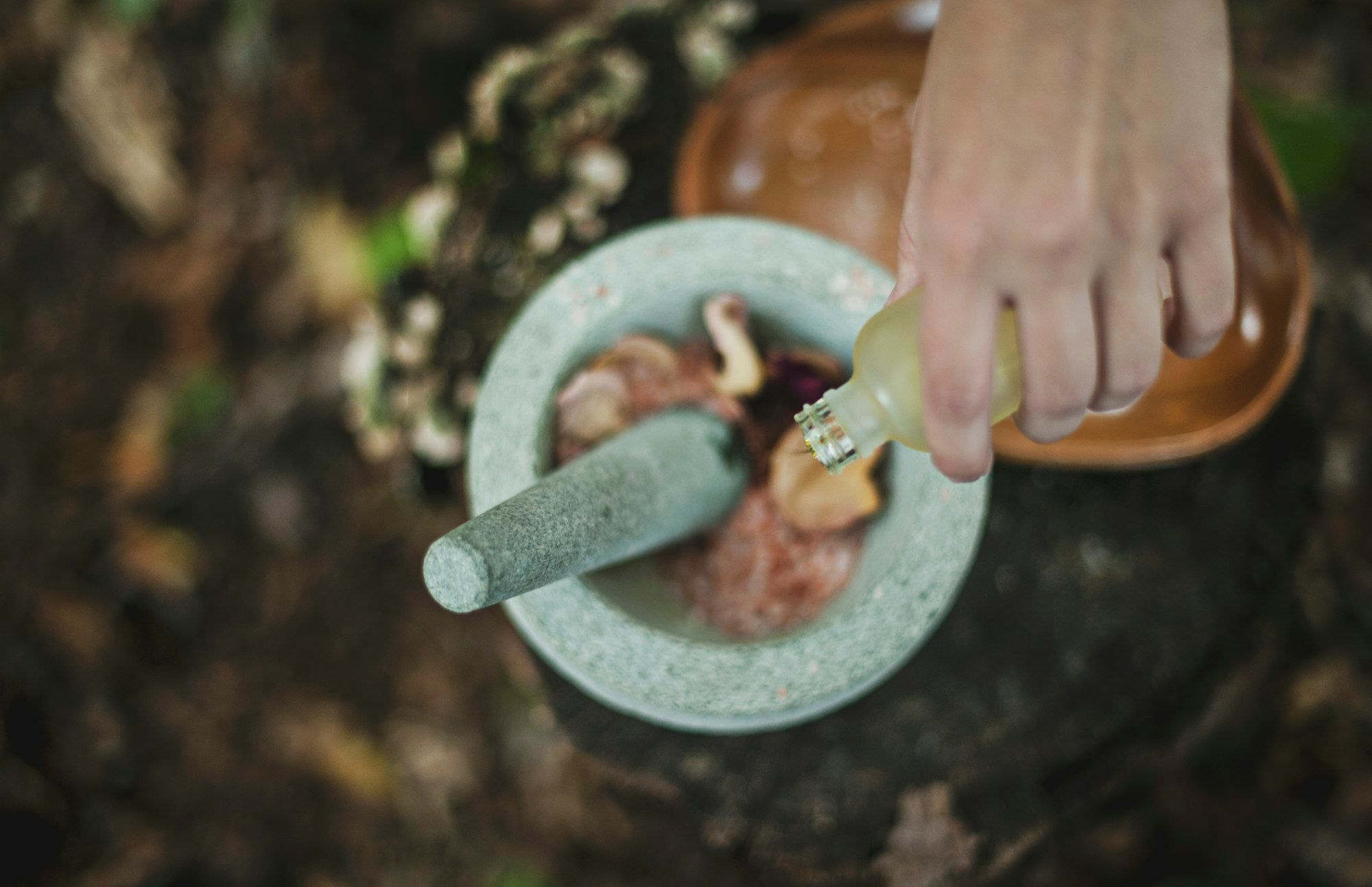 A hand pours herbal liquid into a stone mortar filled with dried herbs and botanicals.