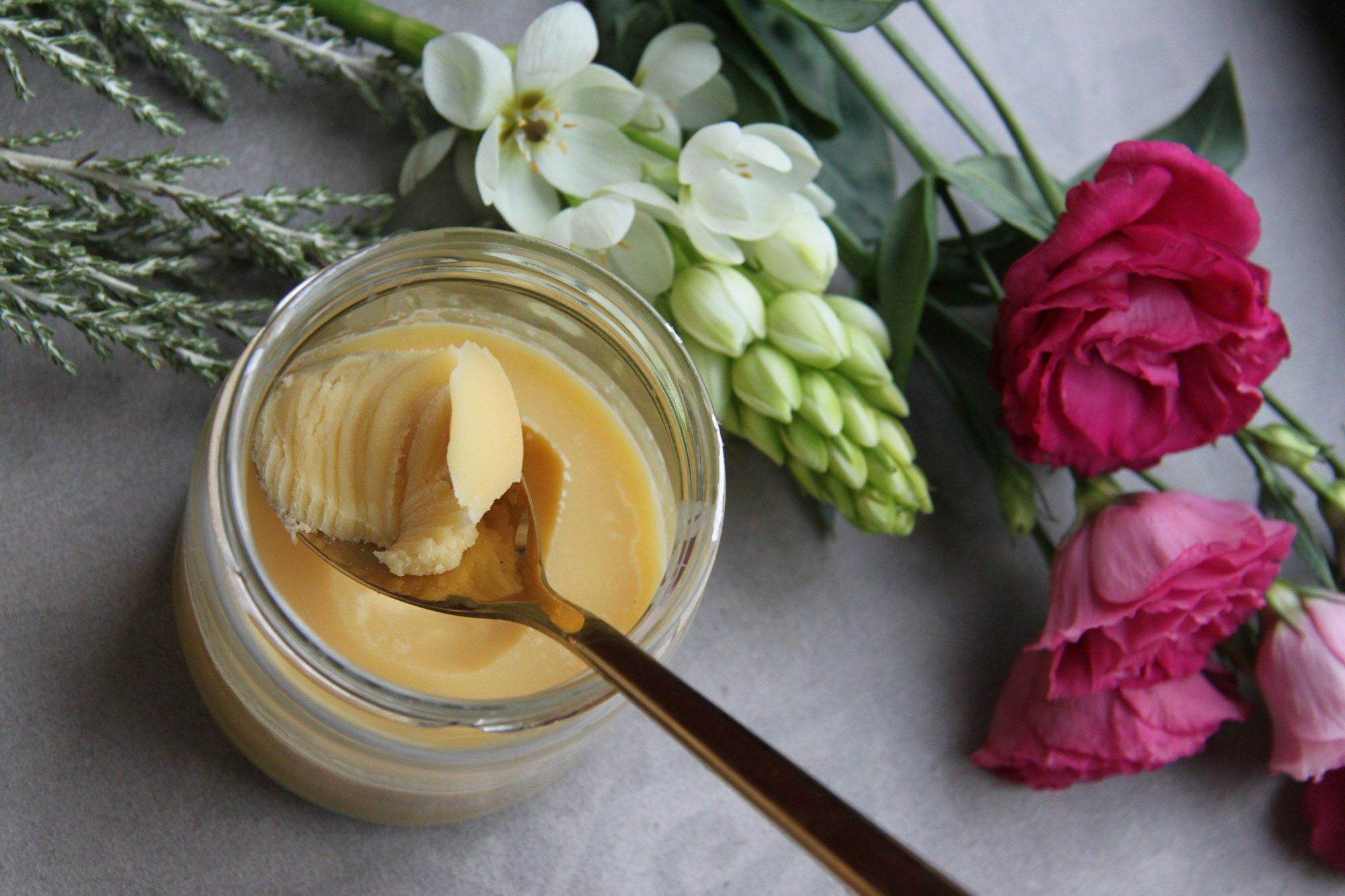 A glass jar of golden homemade ghee with a spoon, set among fresh flowers.