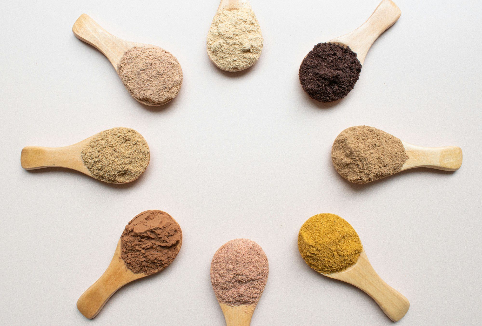 Wooden spoons of different herbal powders arranged in a circle on a white surface.
