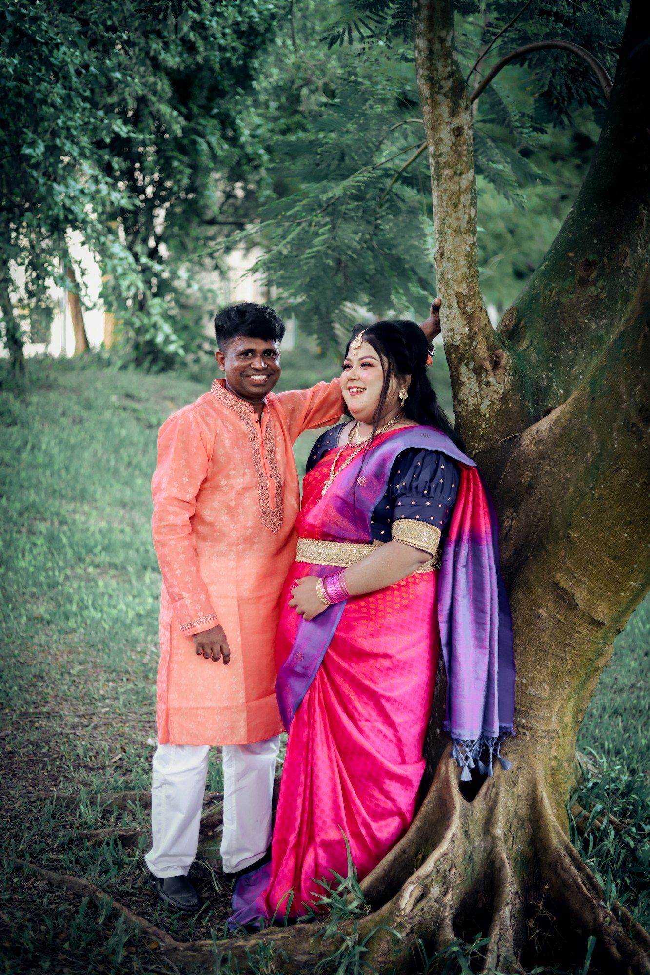An expecting Indian couple in traditional clothes smiling together beside a large tree.