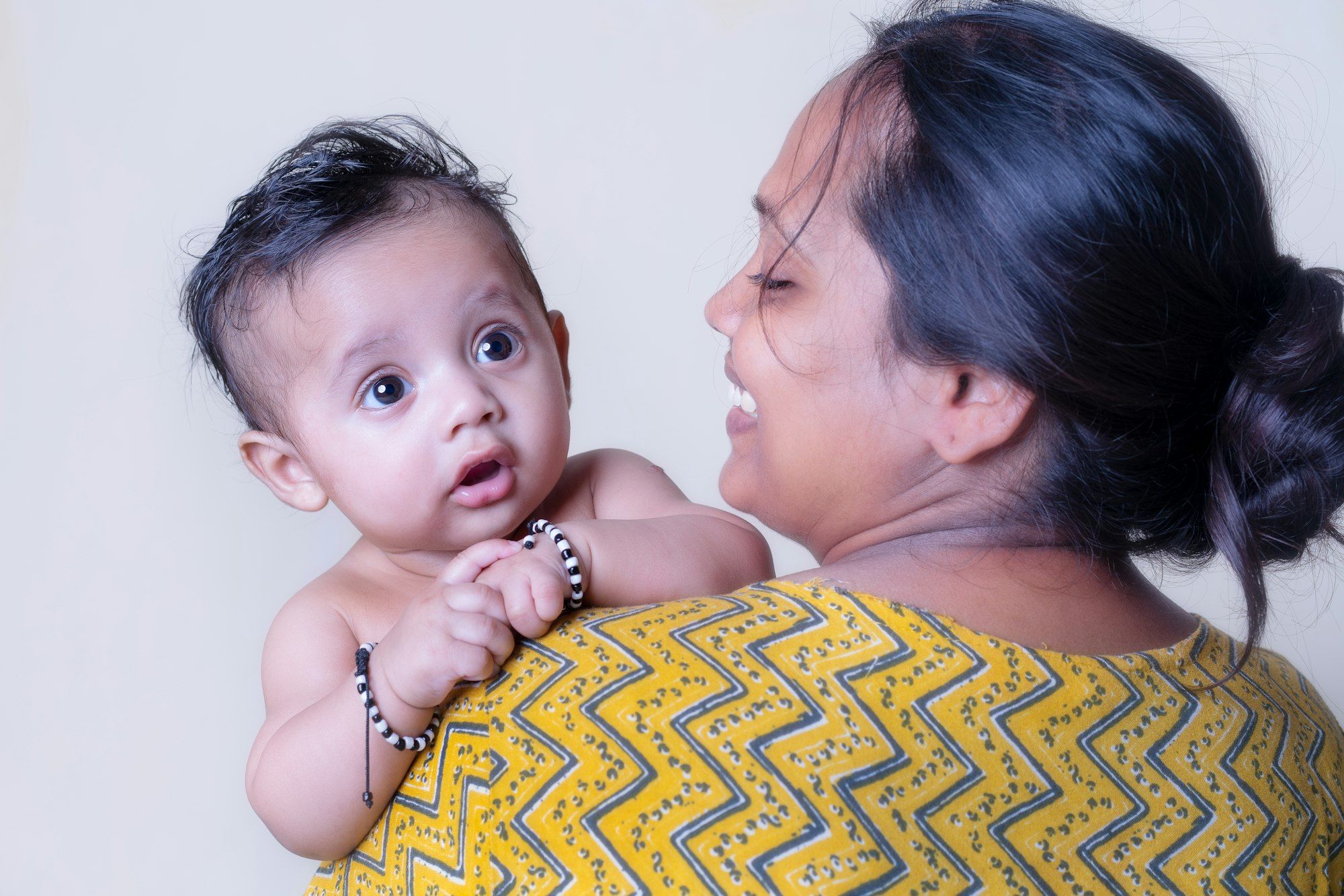 A smiling mother in a yellow printed kurta holding her wide-eyed baby over her shoulder.