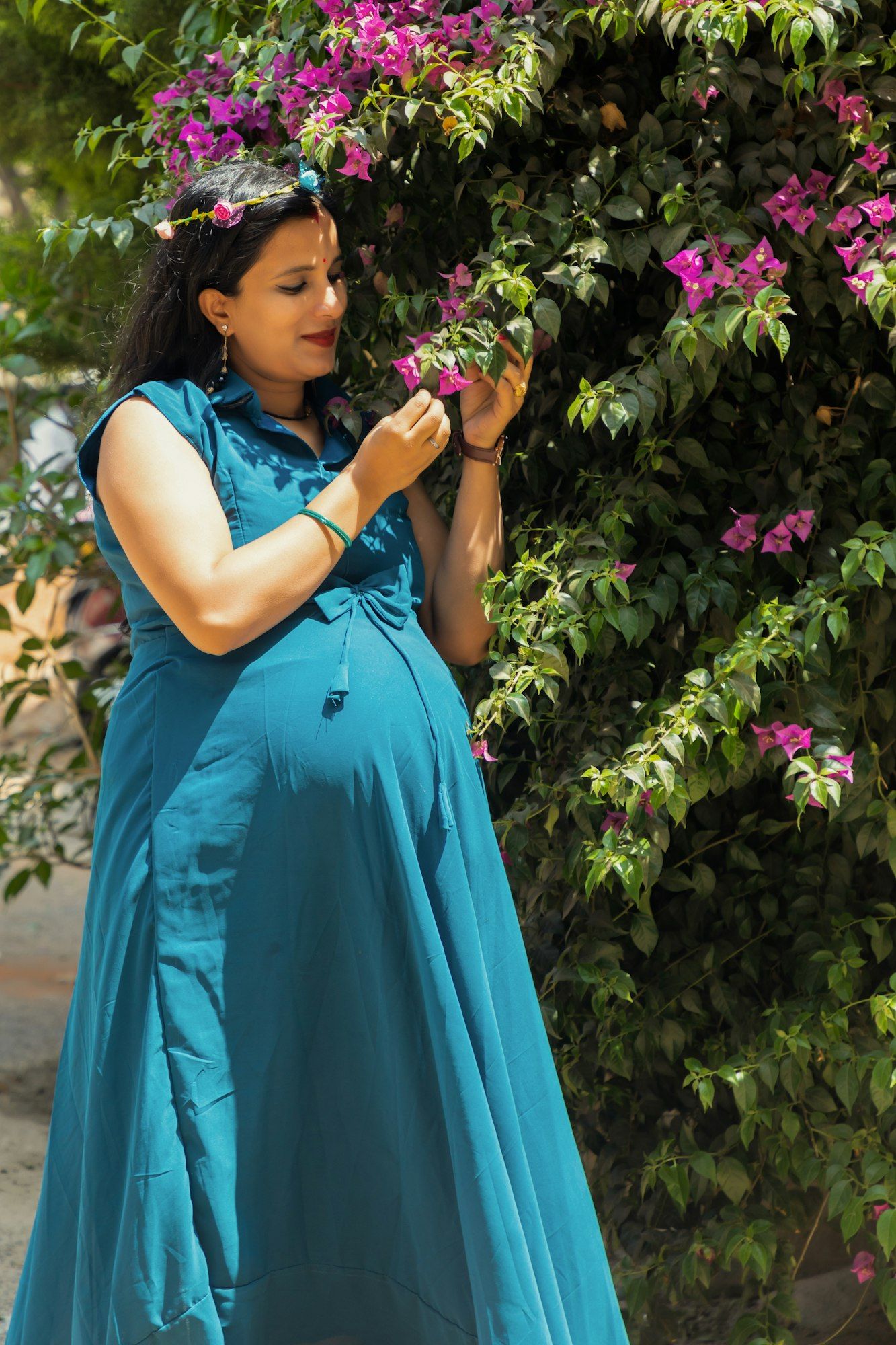 An Indian mother-to-be in a long teal dress smiling softly as she admires pink bougainvillea blossoms in a sunlit garden.