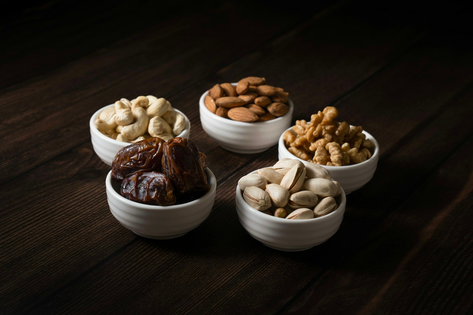 Small white bowls of dates, cashews, almonds, walnuts and pistachios on dark wood.