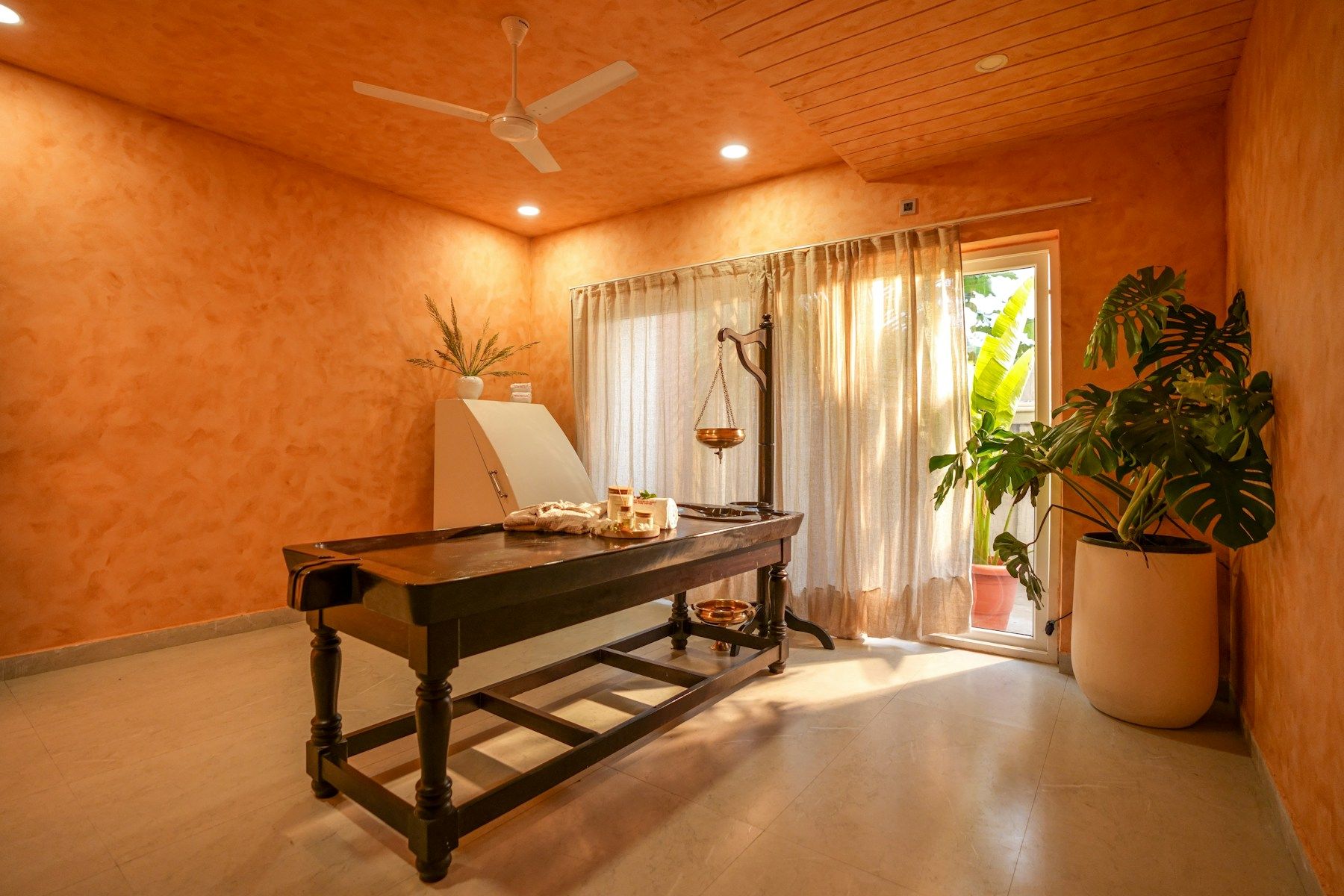 A sunlit treatment room with a wooden therapy table, a hanging oil vessel and potted plants against warm orange walls.